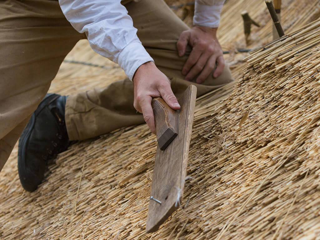 A craftsman is working on laying a thatched roof and shaping the straw with a wooden tool.