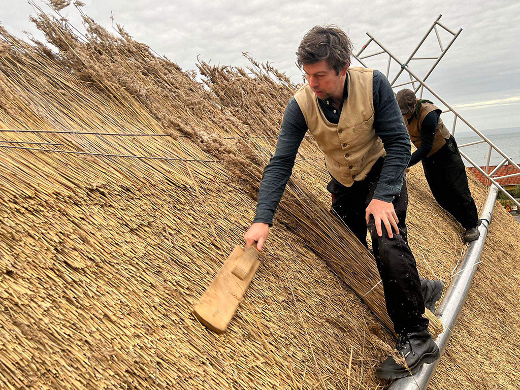 A craftsman is working on laying a thatched roof and shaping the straw with a wooden tool.
